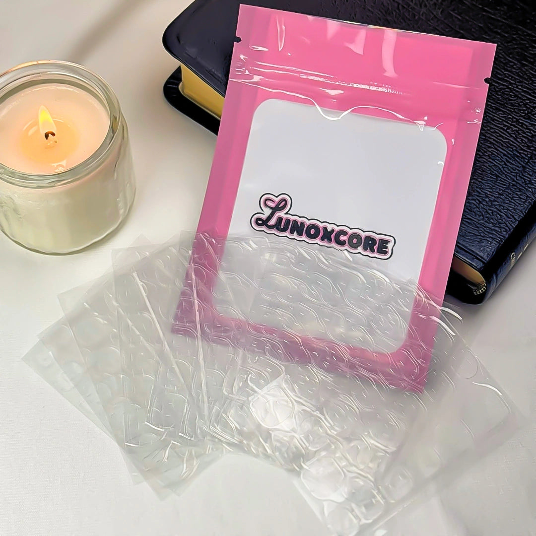 Flat lay of Lunoxcore adhesive tabs kit including pink pouch with Lunoxcore logo, five sheets of clear sticky nail tabs, candle, and book on a white surface — reusable nail stickers for secure press-on wear.