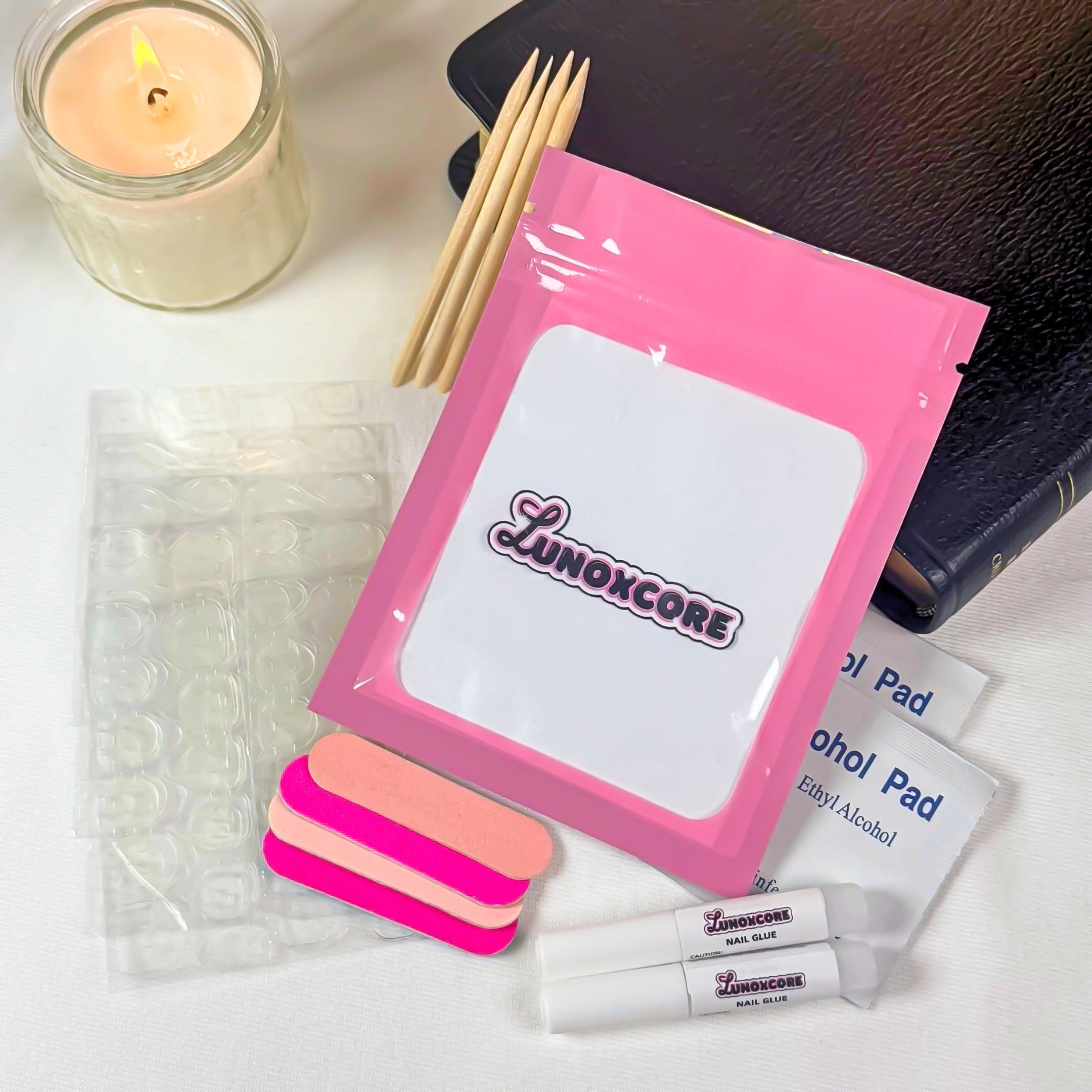 Flat lay of Lunoxcore press-on nail tool kit including pink pouch with Lunoxcore logo, sticky tabs, mini nail files, wooden sticks, alcohol wipes, and nail glue tubes beside a candle and book on a white surface.