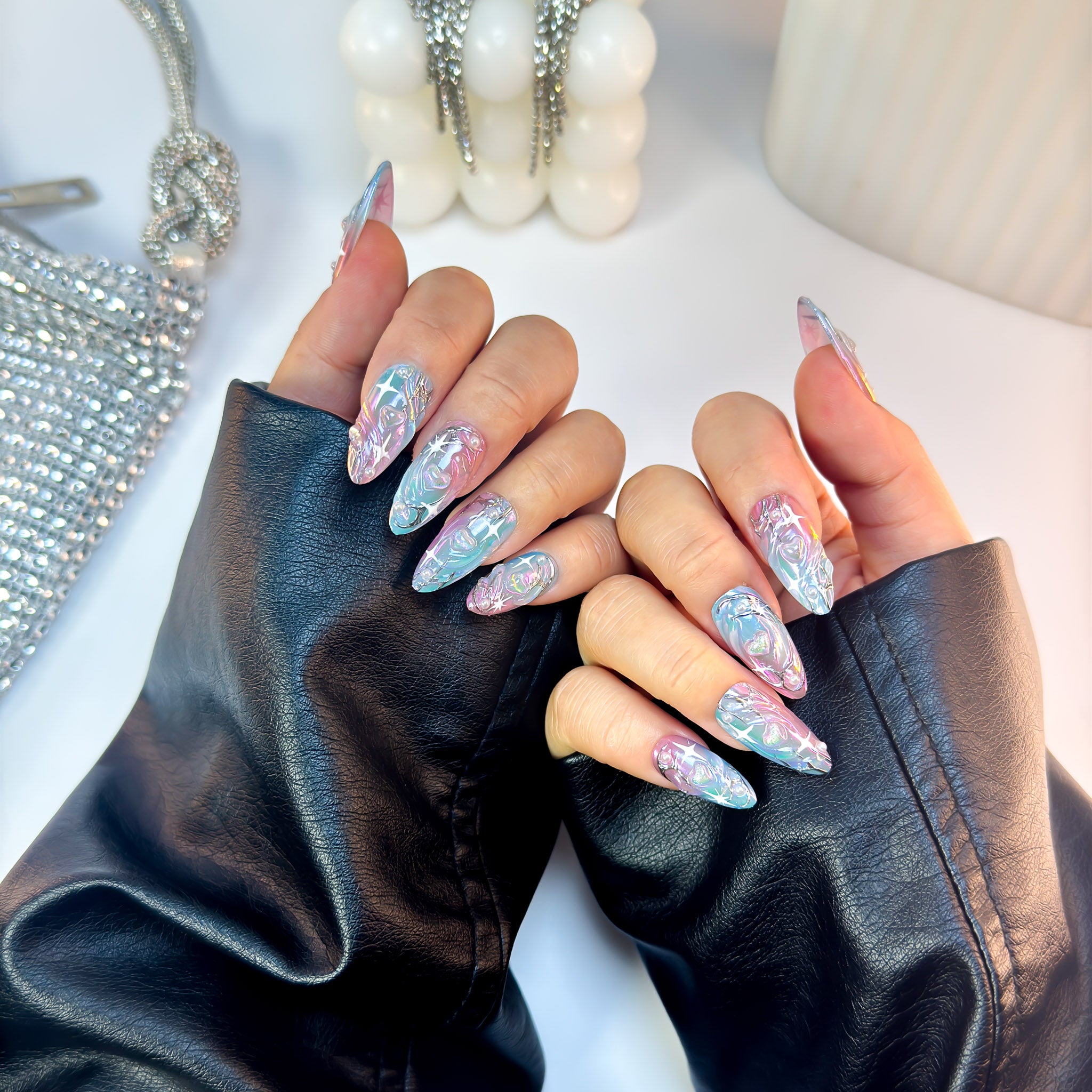 Cosmic press-on nails in reflective pink-blue chrome with metallic contours, displayed with both hands forward wearing black leather sleeves.