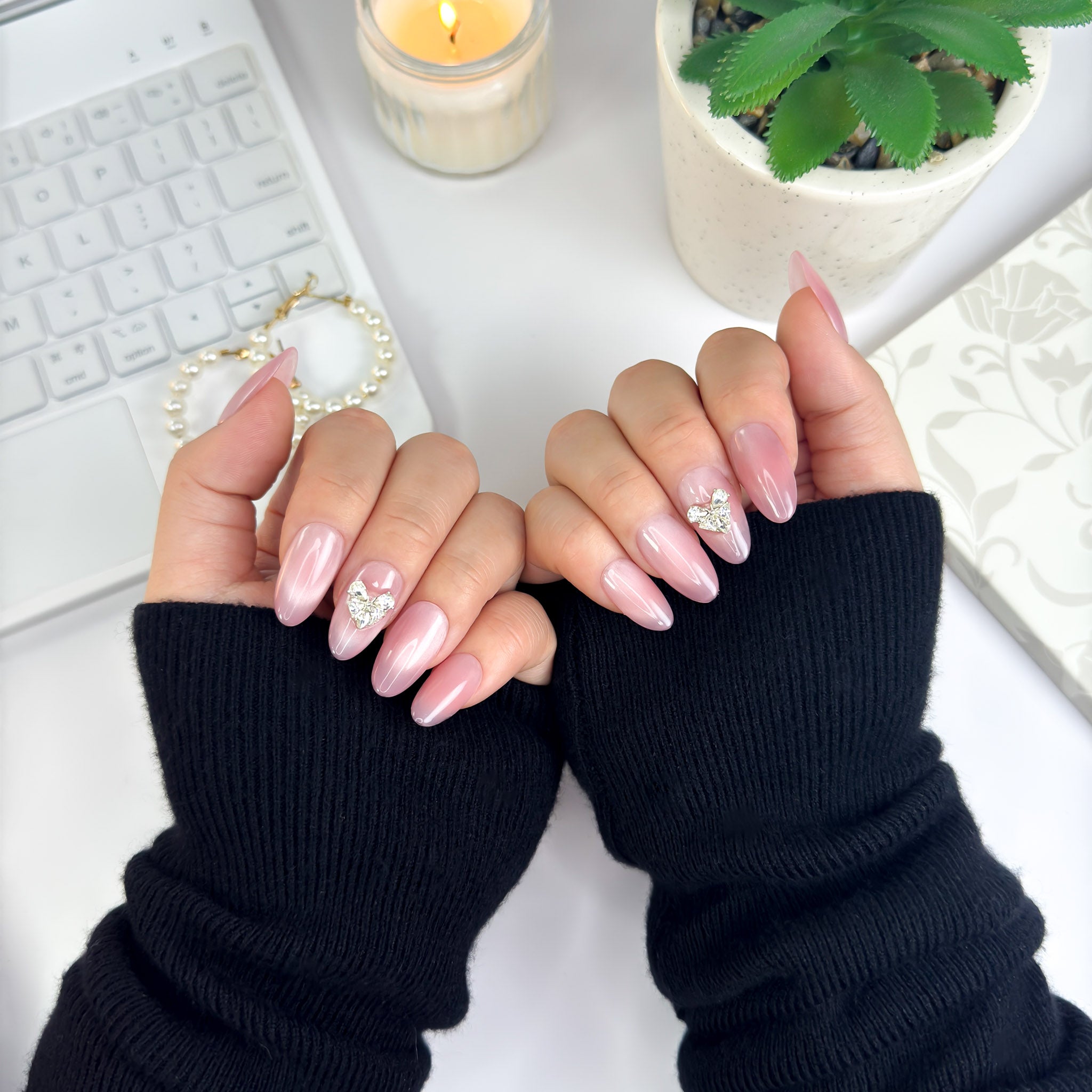 Hands wearing Lunoxcore’s Magnetic Love 3D press-on nails — soft pink chrome finish with heart-shaped crystal accents, shown in a cozy front-facing pose with black sleeves and a candlelit background.