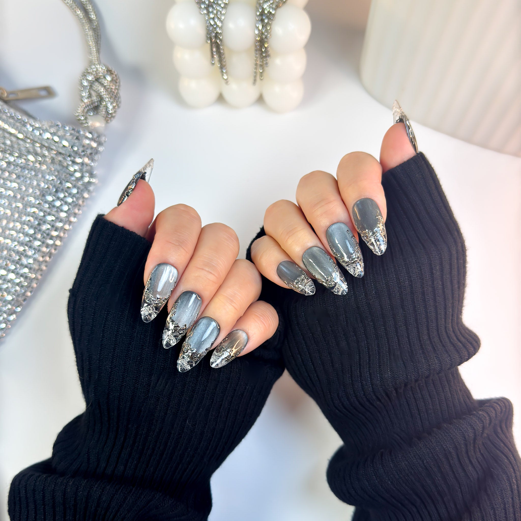 Molten Glass press-on nails in glossy metallic grey with molten silver chrome accents, shown on both hands wearing black ribbed sleeves.