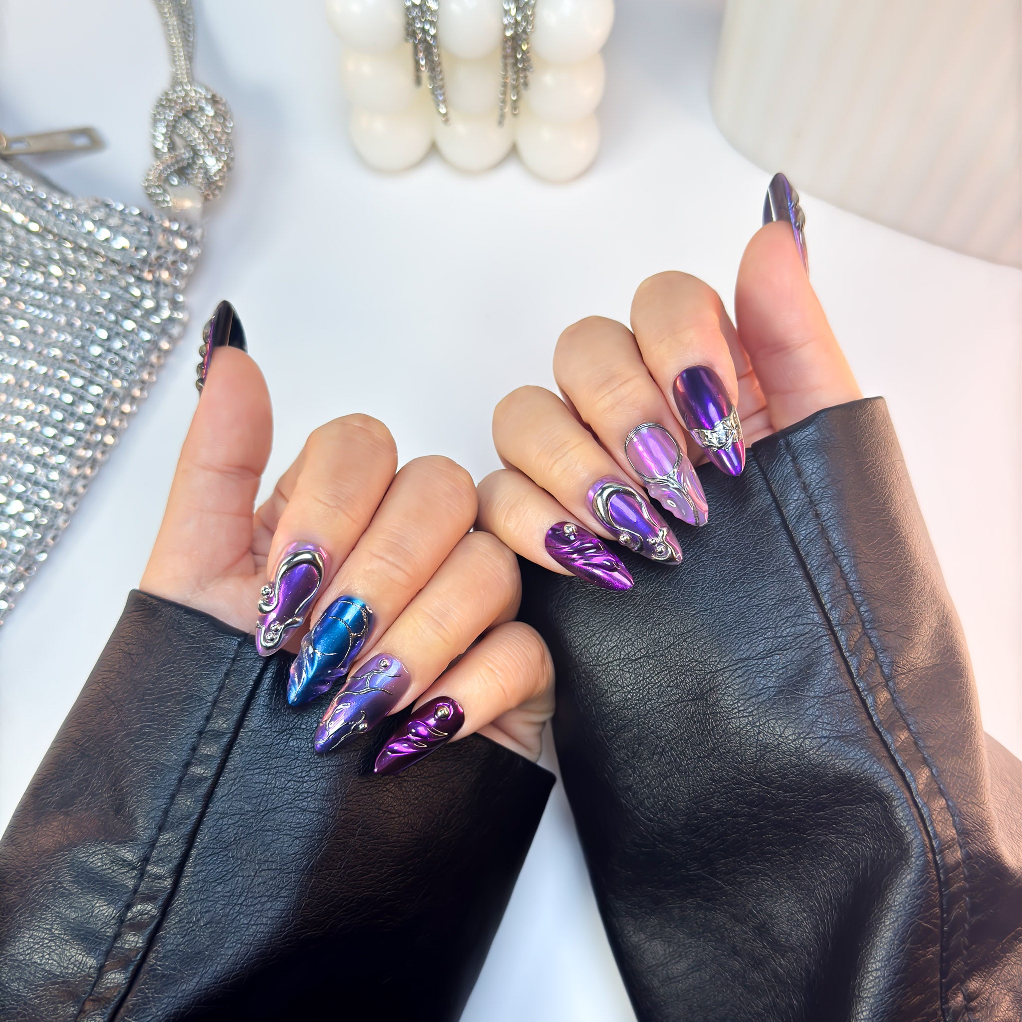 Ultraviolet press-on nails in metallic purple and blue chrome, displayed with both hands together in a front pose wearing black leather sleeves.